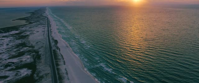 Aerial view of the Florida panhandle coastline at sunset, white sand beach stretching to the horizon with emerald water
